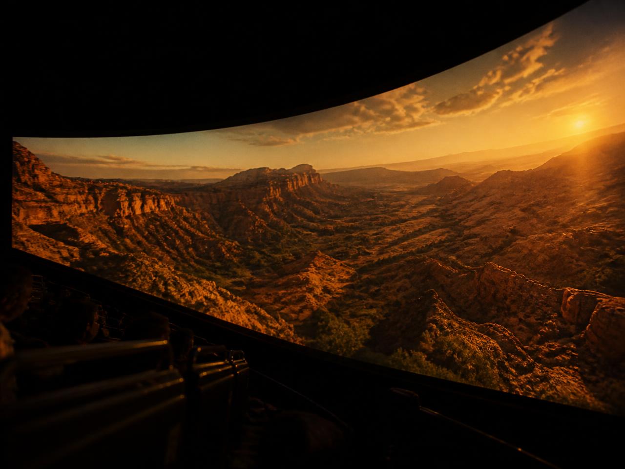 A vast curved cinema screen glowing with aerial footage, filling a dark theater from wall to wall