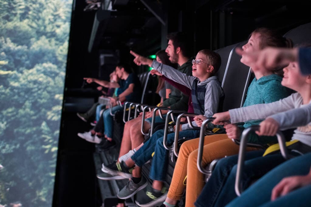 Riders in suspended seats leaning forward, feet dangling, as forest rushes across the curved Take Flight screen