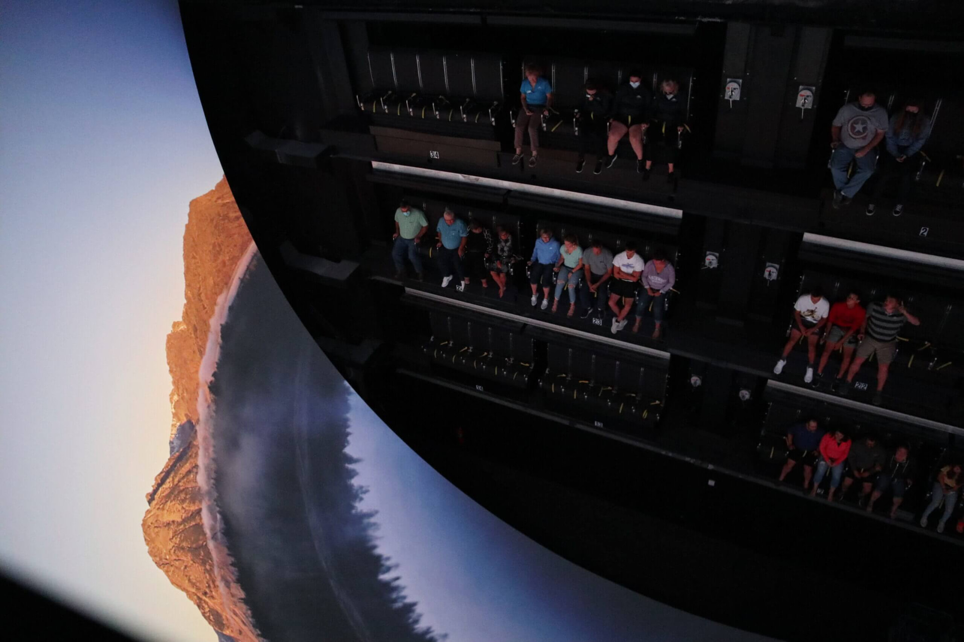 Rows of suspended ride seats filled with guests, seen from above beside the vast curved screen showing a coastline at dawn
